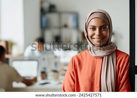 Happy young Muslim female entrepreneur or manager in hijab looking at camera with smile while standing in coworking space