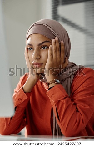 Young troubled businesswoman in hijab touching her face and looking at computer screen while trying to solve technical problem