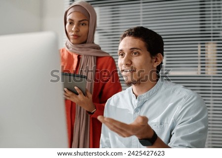 Young confident Muslim male analyst explaining data on computer screen to female colleague in hijab during presentation of project points