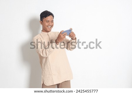 Portrait of young excited Asian muslim man in koko shirt holding mobile phone and playing games on his smartphone. Isolated image on white background