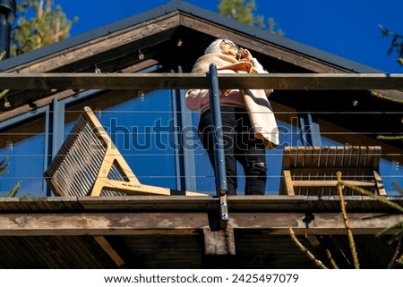 Bottom view of a woman talking on the phone, standing on the terrace of her house in a coniferous forest