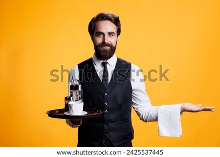 Gracious waiter creating marketing ad in studio, posing with tray in hand and presenting something to left or right sides. Young man working as luxurious butler serving people, elegant personnel. Royalty-Free Stock Photo #2425537445