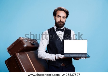 Gracious bellboy holds laptop with white screen in studio, presenting new blank copyspace technology as hotel concierge. Young man working as doorkeeper porter, modern hospitality industry. Royalty-Free Stock Photo #2425537527