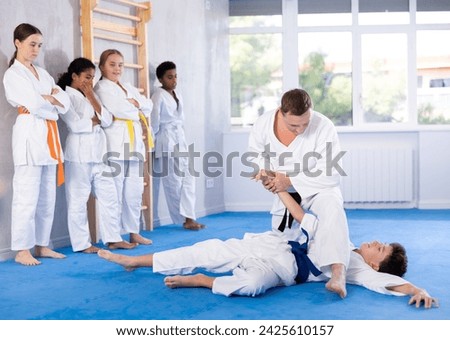 Experienced judo master conducting classes for teenage beginners, demonstrating armbar technique in sparring with student to group of teenagers standing by wall in training room.. Royalty-Free Stock Photo #2425610157
