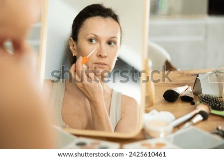 Careful middle-aged lady doing makeup with concealer while seated at a table in her house Royalty-Free Stock Photo #2425610461