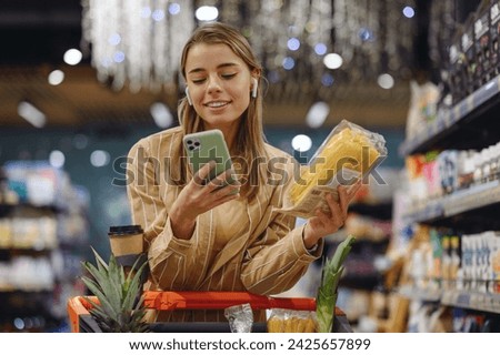 Young woman wears casual clothes earphones listen music use mobile cell phone hold pasta shopping at supermaket store grocery shop buy with cart choose products in hypermarket. Purchasing food concept