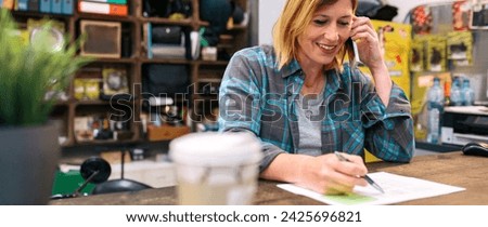 Portrait of smiling blonde young woman receiving order by phone for ecommerce shop. Happy woman with freckles wearing a plaid shirt and talking with cellphone while filling selling form in a store.