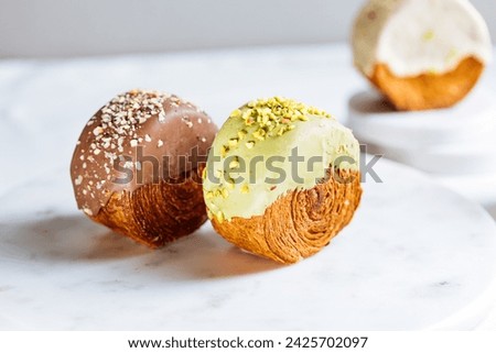 Fusion croissant called Cromboloni invented in New York. Round puff pastry filled with cream, and glazed with white, milk chocolate or pistachio. Crunchy and soft in the same time. Bright background.  Royalty-Free Stock Photo #2425702097