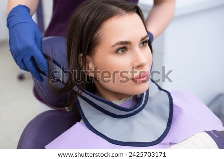 The dental assistant outfits the patient with a protective neck shield before commencing the tooth X-ray procedure.  Royalty-Free Stock Photo #2425703171