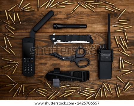 Close-up on a wooden background. Satellite phone with a large antenna, radio station, tactical knife, flashlight, atraumatic scissors, and hemostatic tourniquet. Frame of cartridges