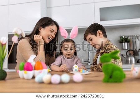 Cute little children wearing funny bunny ears headbands embracing and kissing young happy mother while painting Easter eggs together, sitting at table in kitchen, selective focus