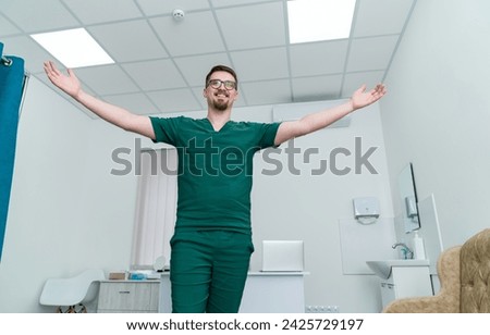 Male doctor in green uniform stands in room with his arms outstretched.