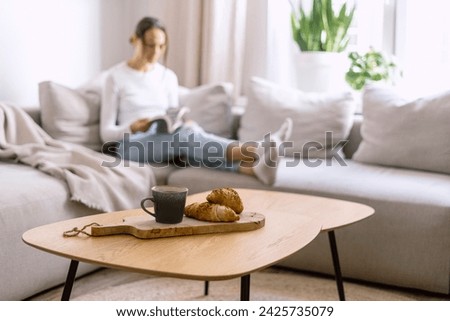 Selective focus on fresh croissants and cup with coffee on wooden table against blurred background with woman reading book in modern living room. Tasty breakfast in morning. Spending time at home. Royalty-Free Stock Photo #2425735079
