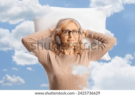 Middle aged woman with headphones resting on a pillow among clouds on a blue sky