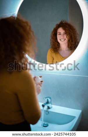 Beautiful young woman with lush curly hair standing in front of bathroom mirror and smiling at her reflection.