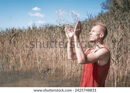 A man wearing a muscle shirt stands on the shore of a lake and has scooped water into the air. Royalty-Free Stock Photo #2425748831
