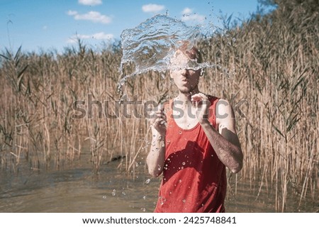 A man wearing a muscle shirt stands on the shore of a lake and has scooped water into the air. Royalty-Free Stock Photo #2425748841