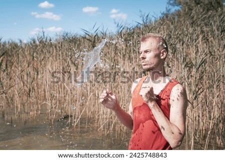 A man wearing a muscle shirt stands on the shore of a lake and has scooped water into the air. Royalty-Free Stock Photo #2425748843