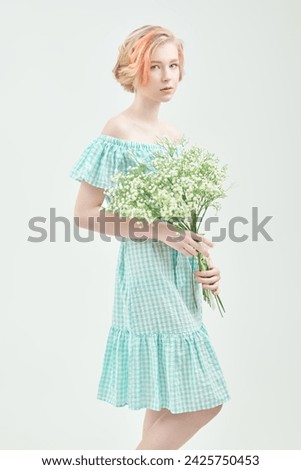 A cute blonde teen girl with a short haircut in a summer sundress and a pearl necklace poses against a white wall with a bunch of flowers. Delicate spring-summer look. Beauty, fashion.