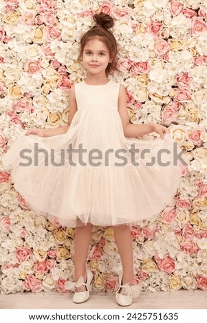 A cheerful emotional girl in a light white dress poses against a floral background. Children's fashion. Full-length portrait. 