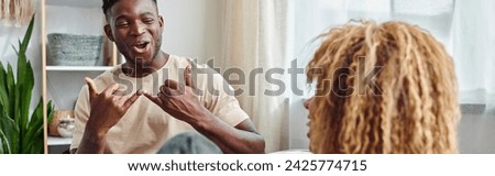 joyful black man smiling while communicating with sign language with girlfriend at home, banner