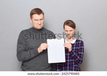 Studio portrait of adult couple holding together white blank paper sheet with place for your text, both are angry, disgruntled and frowning, with negative expressions, standing over gray background