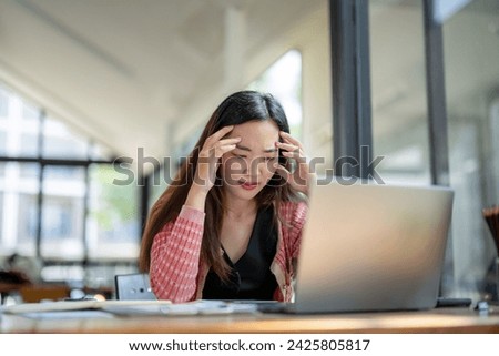 Young Asian businesswoman feeling stressed while working on a laptop in a modern office setting.
