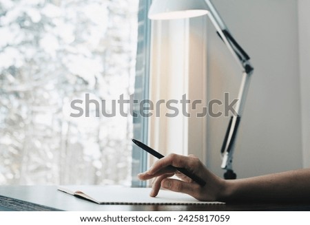 Photo of a hand at a desk. A woman's hand holds a pen. Notepad on the table. Workplace with a view of the forest.
