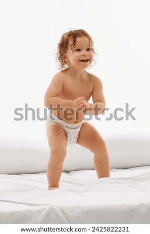 Carefree and playtime. Adoring baby with radiant smile stands in diaper on comfy bed against white background. Concept of beauty, childhood, motherhood, life, birth. Copy space for ad