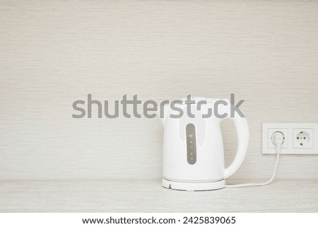 New white plastic electric kettle with plug in wall outlet socket on stone table top at home kitchen. Closeup. Empty place for text on beige wallpaper background. Front view.
