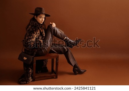 Fashionable confident woman wearing hat, trendy long faux fur leopard print coat, leather pants, pointed toe boots, with shoulder bag, posing on brown background. Full-length studio fashion portrait 