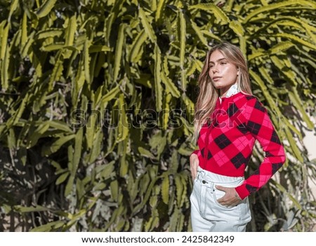 Hight contrast e-commerce in nature: Beautiful, slim blonde hair model posing in front of a rustic house surrounded by cacti in a green environment. Wearing a red sweater with jeans and black shoes
