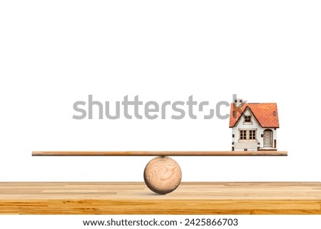 White house with a red roof. The house is sitting on a wooden seesaw, which is balanced on a wooden table. Financial realestate business concept