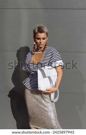 Spectacular blonde girl in sequin skirt and t-shirt posing over grey wall. Close-up outdoor portrait of glamour european woman holding a white leather shopper bag
