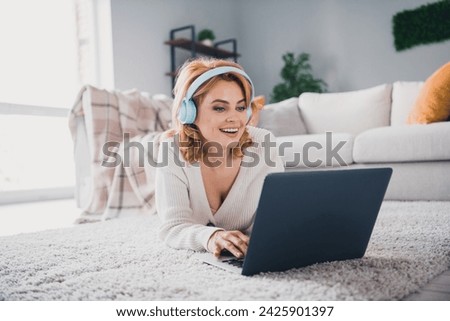 Photo of charming cheerful lady laying carpet floor listen music headphones use wireless laptop modern interior flat indoors