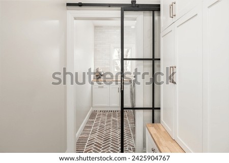 Elegant Laundry Room with Neutral Color Palette and Transitional Design. Black Framed Sliding Glass Barn Door with Mudroom Storage and Brick Floor. Royalty-Free Stock Photo #2425904767