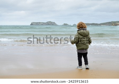 Horizontal photograph with copy space of an unrecognizable boy standing in front of the sea on a beach with his coat on on a cold winter day. Freedom, carefree, nature