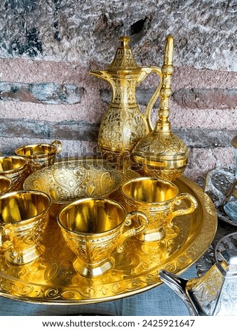Vintage golden tea set with intricate designs on tray against rustic stone wall. Traditional ornate metalware concept for interior design and decoration. Royalty-Free Stock Photo #2425921647