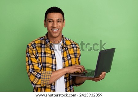 Portrait of handsome young smiling man in plaid shirt using netbook to control task managing people isolated on green color background