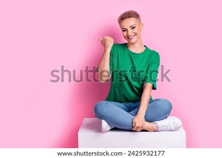 Full size photo of optimistic cute girl sit on white cube indicating at proposition empty space isolated on pink color background