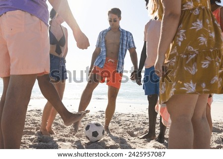 Diverse friends, who are young adults, enjoy a casual game of soccer on the beach. Sunlight enhances the carefree outdoor atmosphere as they play.