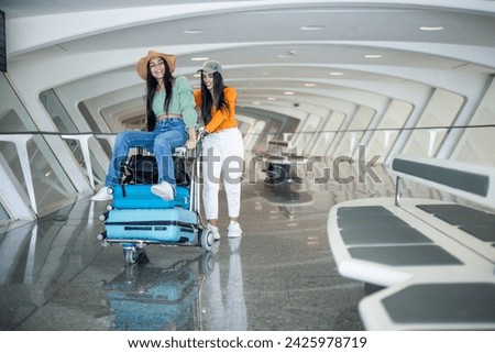 couple of two multicultural girls at the airport sitting on the luggage cart walking having fun Royalty-Free Stock Photo #2425978719