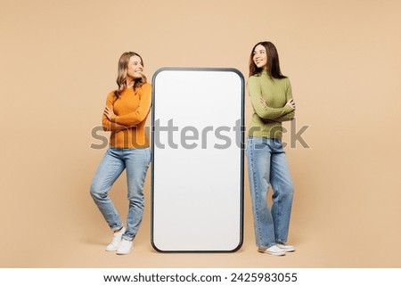 Full body young happy friends two women they wear orange green shirt casual clothes together stand near big huge blank screen mobile cell phone smartphone with area isolated on plain beige background