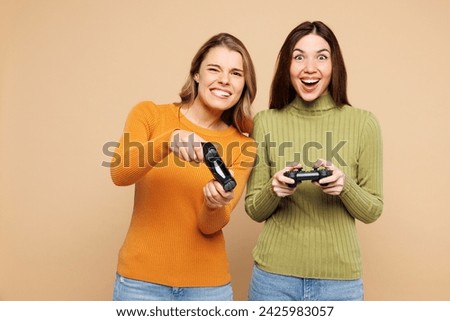 Young friends two women they wear orange green shirt casual clothes together hold in hand play pc game with joystick console isolated on plain pastel light beige background studio. Lifestyle concept