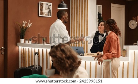 Happy guests on business trip and professional resort employee chatting in cozy stylish hotel lobby. Friendly receptionist handing company coworkers room access key card during check in process.