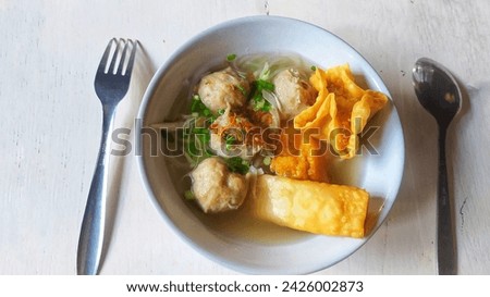 This is an Indonesian food called bakso bakwan which consists of meatballs, vermicelli noodles, fried tofu, dumplings doused in beef broth served in a bowl and accompanied by a spoon and fork. Royalty-Free Stock Photo #2426002873