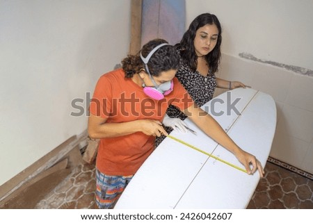 Top view photo of a worker and assistant measuring the surface of a surfboard in a repair shop