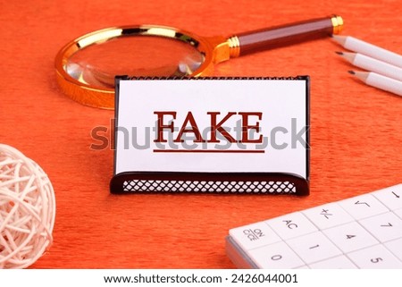 FAKE word written on a white business card on a stand on an orange background, next to a magnifying glass, a calculator and pencils