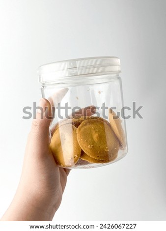 Hand holding round plain sweet bakery food biscuit treats inside clear plastic glass jar container. Object photography isolated on white studio background.