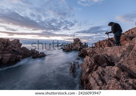 A man capturing photos of the Mediterranean Sea with his camera and tripod at sunrise. Costa Brava, Cala Rosamar